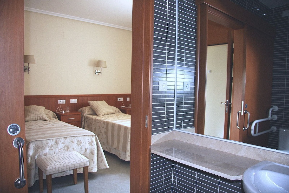 Une chambre d’hôtel avec deux lits, une salle de bain équipée d’un lavabo et d’un miroir, et une fenêtre avec des stores.