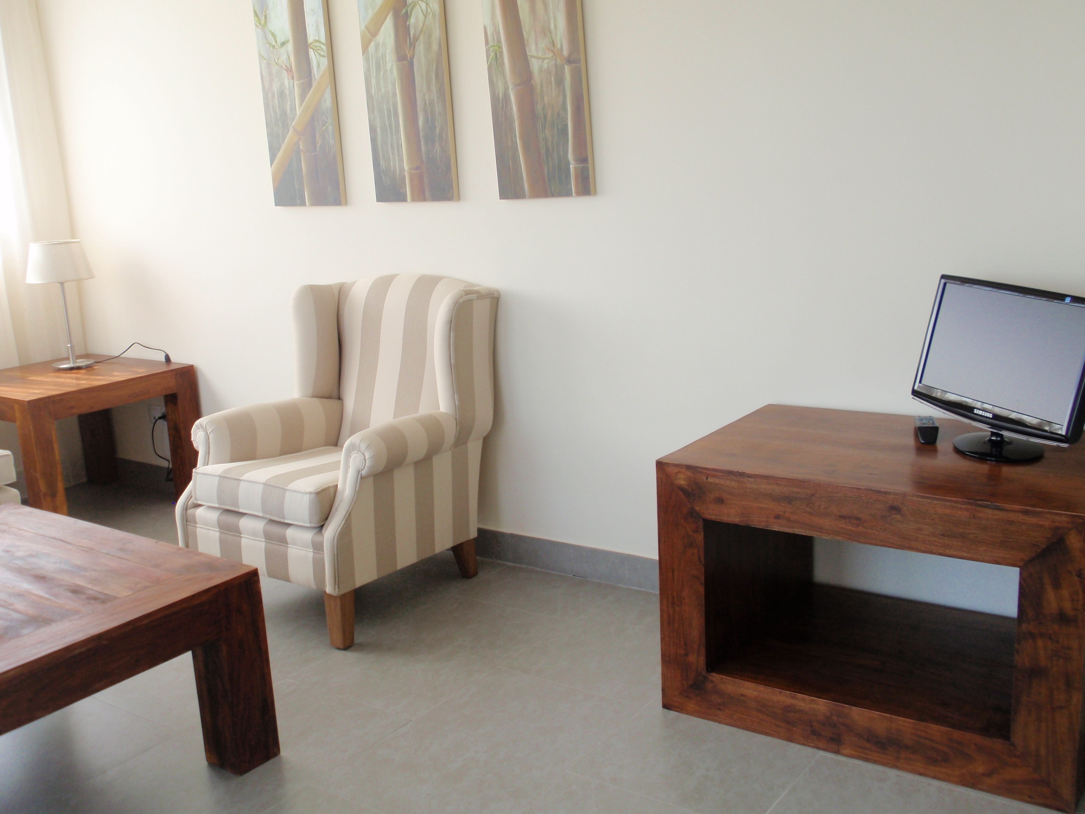 Una habitación con un sillón a rayas, una mesa de madera, un televisor sobre un mueble de madera y una lámpara en una mesita auxiliar.