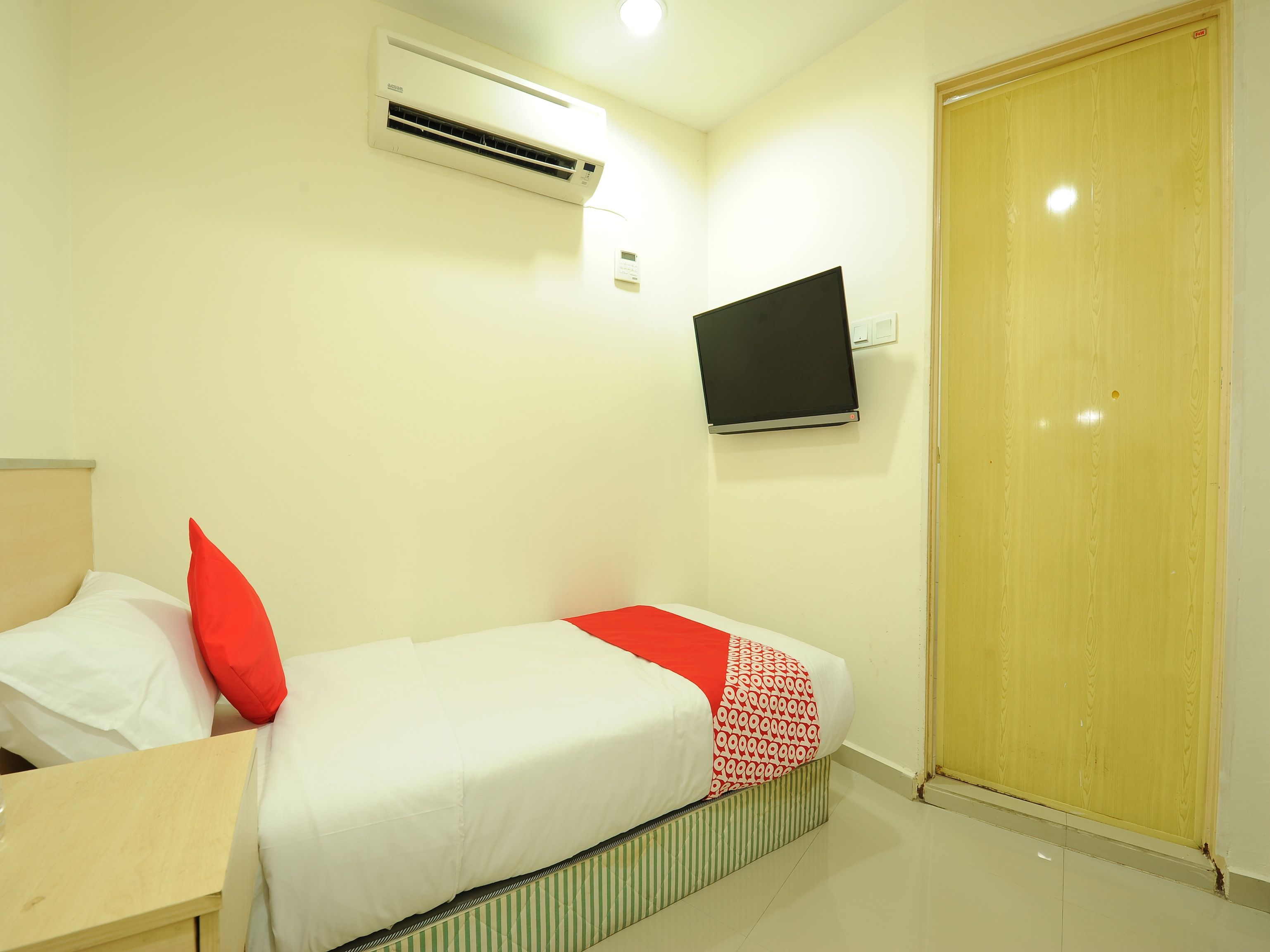 A hotel room with a bed, a TV mounted on the wall, an air conditioning unit, and a wooden door.