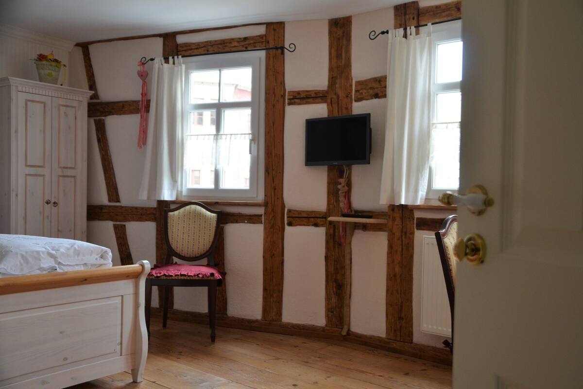 Un dormitorio con techo de madera, una cama, una silla, un televisor y un ventanal con cortinas.