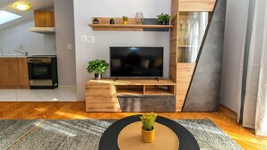 Una sala de estar moderna con un televisor de pantalla plana, muebles de madera, una cocina al fondo y un suelo con estampado tipo espiga.