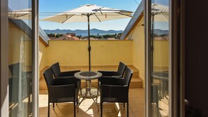Un balcón con dos sillas de mimbre, una mesa redonda, un paraguas y vistas a montañas y edificios lejanos.