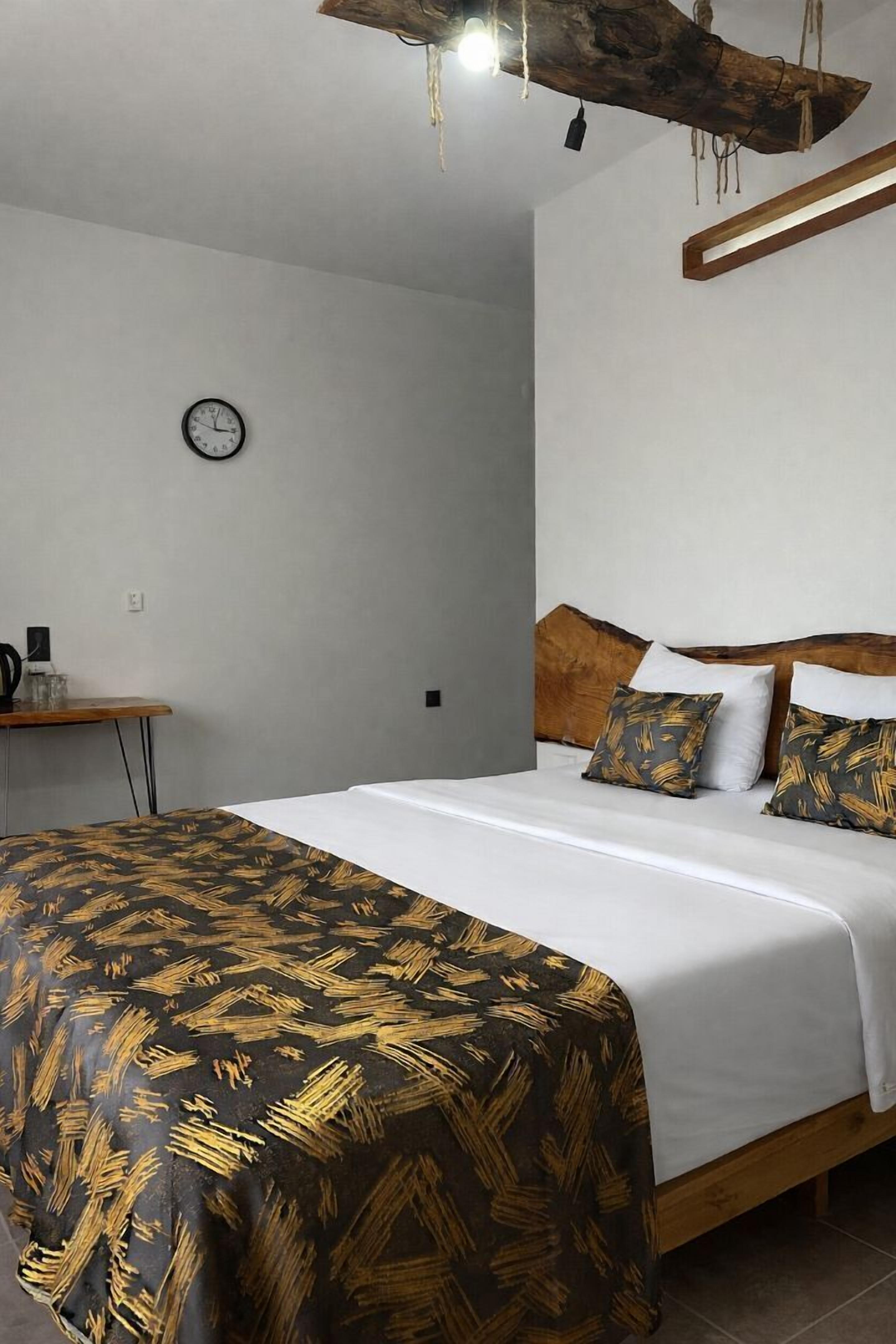 Un dormitorio con cabecera de madera, cama con colcha estampada y una mesita de noche con hervidor, además de un reloj de pared.