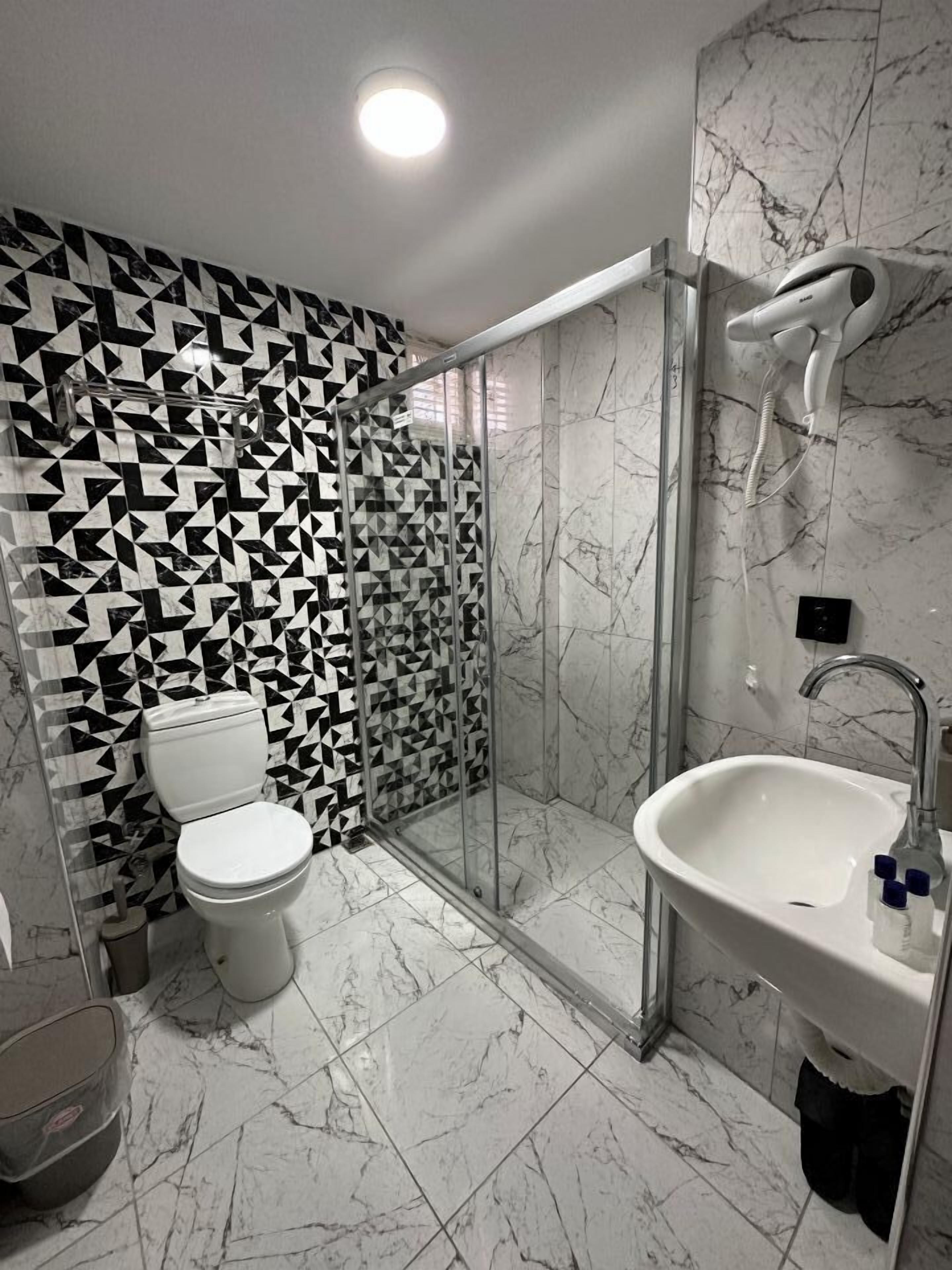 Une salle de bain moderne avec un mur orné d’un motif géométrique, une douche en verre, des toilettes blanches et un lavabo avec un miroir.
