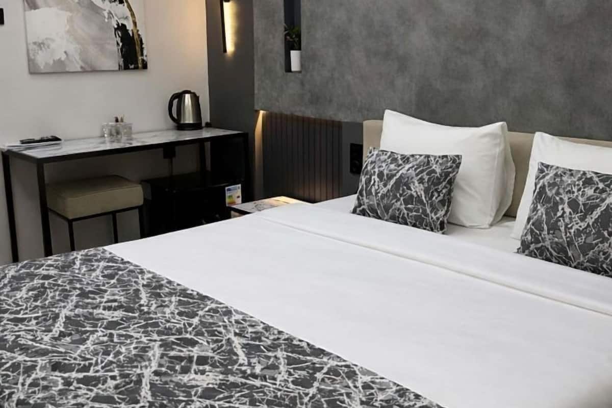 A modern hotel room with a large bed, a desk with a chair, a coffee maker, and a painting on the wall.