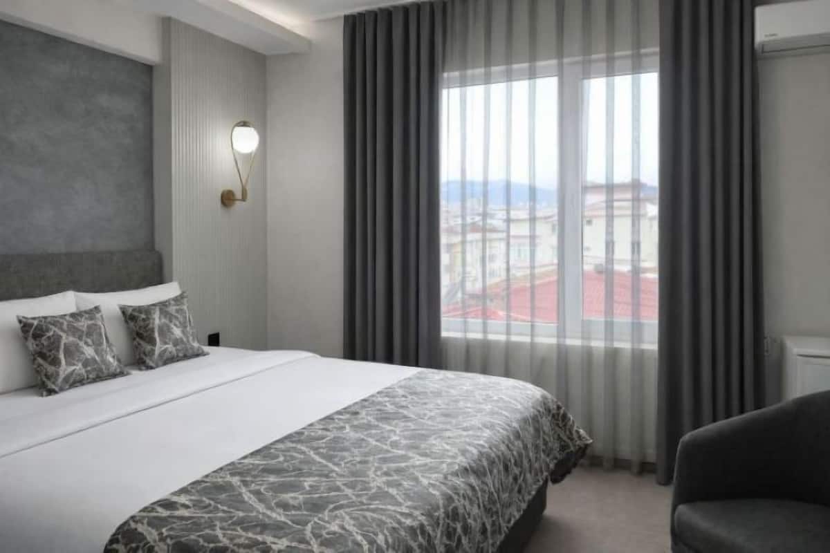 A hotel room with a large bed, grey curtains, a city view, and a modern light fixture.