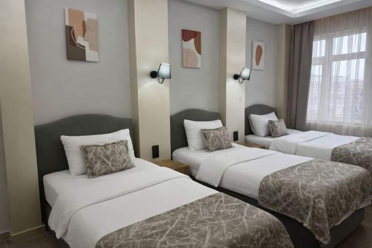 A hotel room with three beds arranged in a row, each with a grey headboard and white bedding. There are abstract wall art pieces above each bed and a large window with curtains on the right.