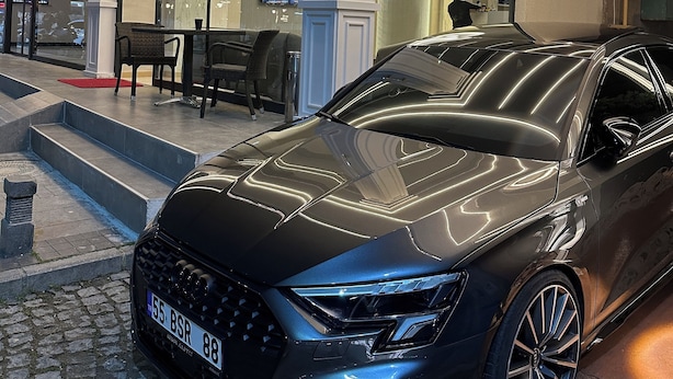 Une voiture de luxe noire est garée devant un hôtel au design moderne, qui se distingue par son entrée imposante et un espace salon visible à l’intérieur.