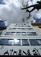 Egy modern épület, amelynek homlokzatán az „ATHENS 21” felirat látható, szabályos elrendezésű ablakokkal és egy fáraggal, amely a bejárat fölé nyúlik.