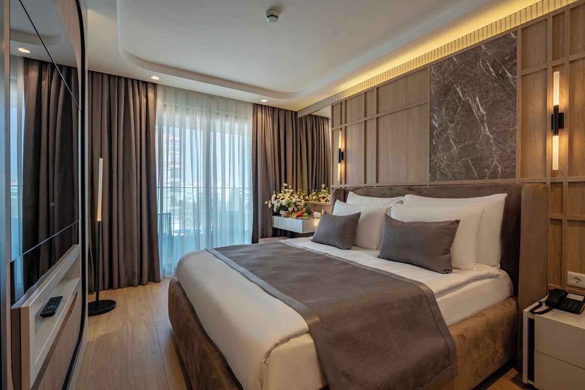 A modern hotel room with a large bed, a bedside table, a nightstand, and a view of the city through large windows.