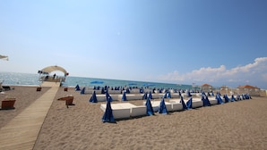 A beach with rows of blue umbrellas and white lounge chairs, a wooden walkway, and a clear view of the sea. - Awen Lara Hotel (Antalya)