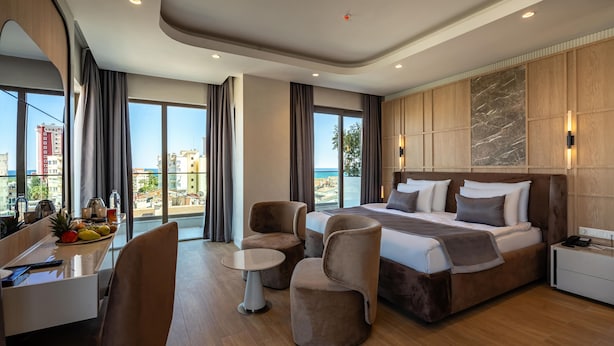 A modern hotel room with a large bed, a desk with a chair, a small table, and a view of the city and sea. - Awen Lara Hotel (Antalya)