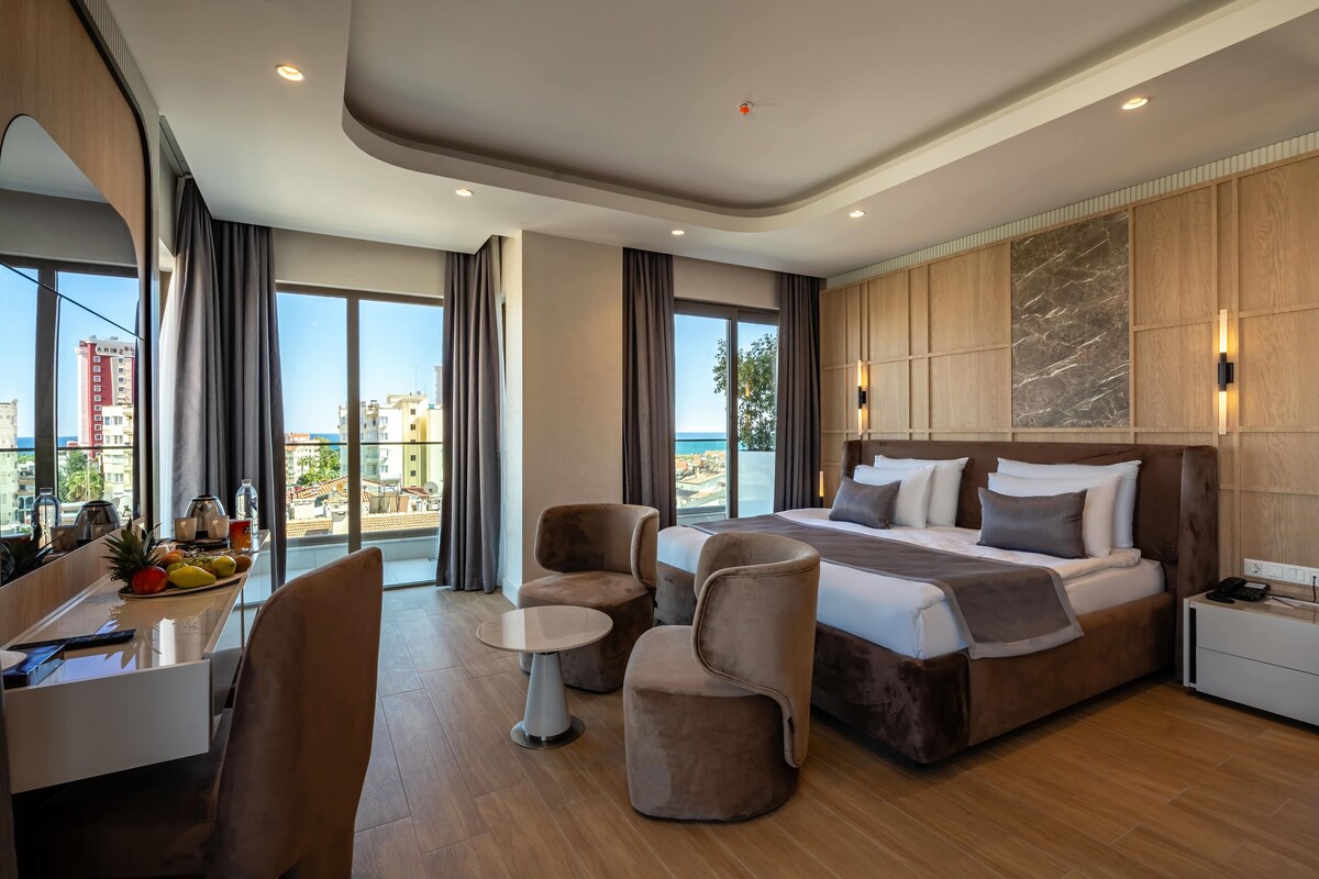 A modern hotel room with a large bed, a desk with a chair, a small table, and a view of the city and sea.