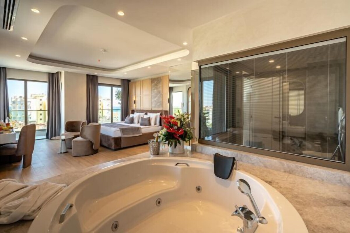 A modern hotel room with a large bathtub, a sofa, a dining area, and a large window with a view.