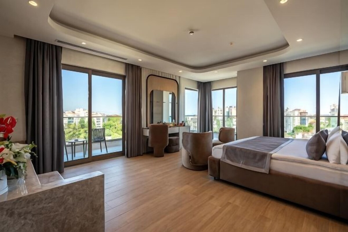 A modern hotel room with a large bed, a seating area, a desk, and large windows offering a city view.