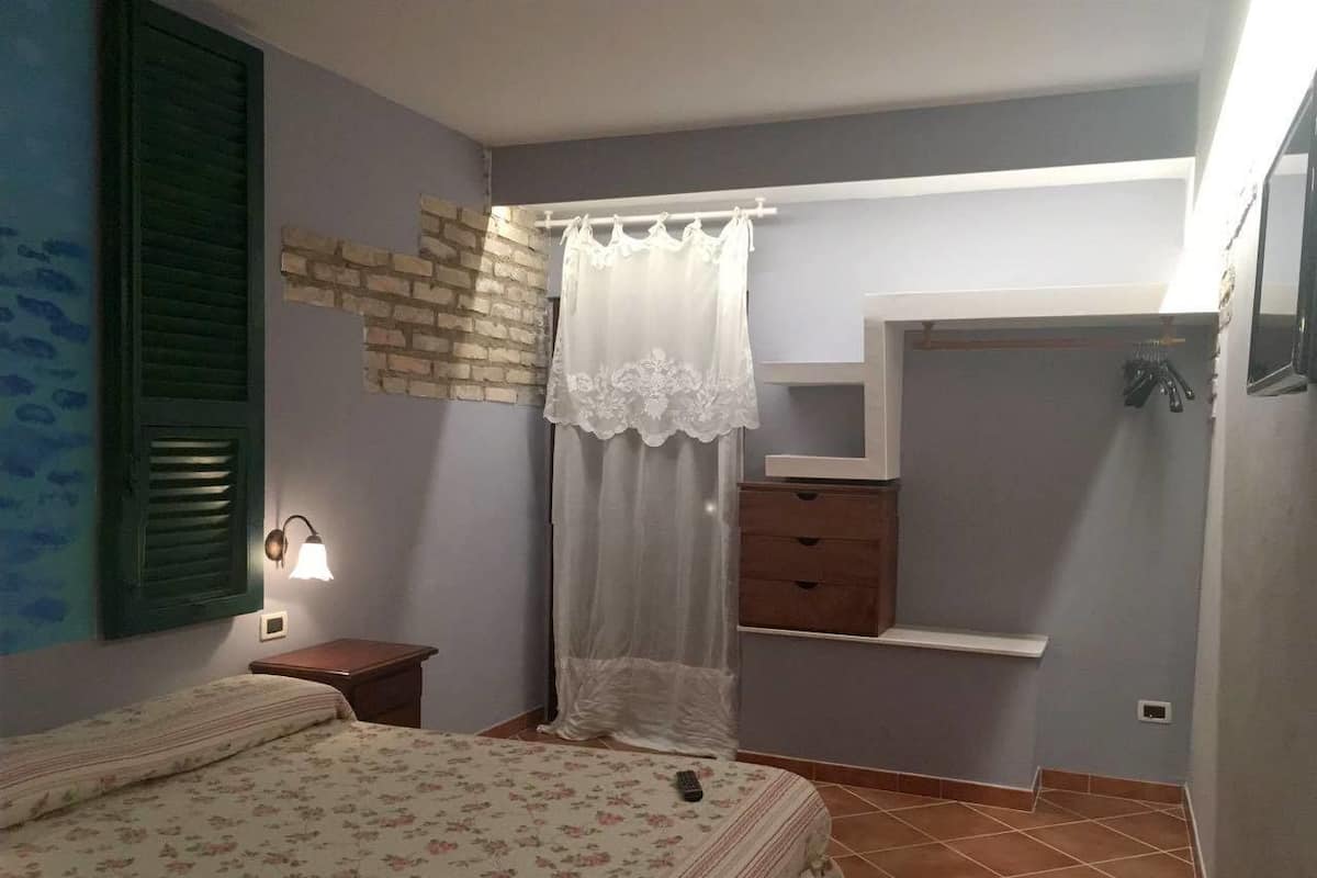 Quarto com cama, mesa de cabeceira, cômoda, janela com persianas e prateleira na parede.