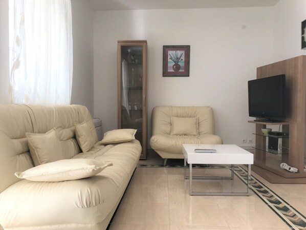 A living room with a beige sofa, a matching armchair, a white coffee table, a television on a wooden stand, and a framed picture on the wall.