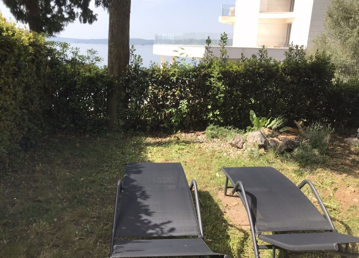 Duas espreguiçadeiras numa área ajardinada, com vista para um corpo de água e um edifício ao fundo.