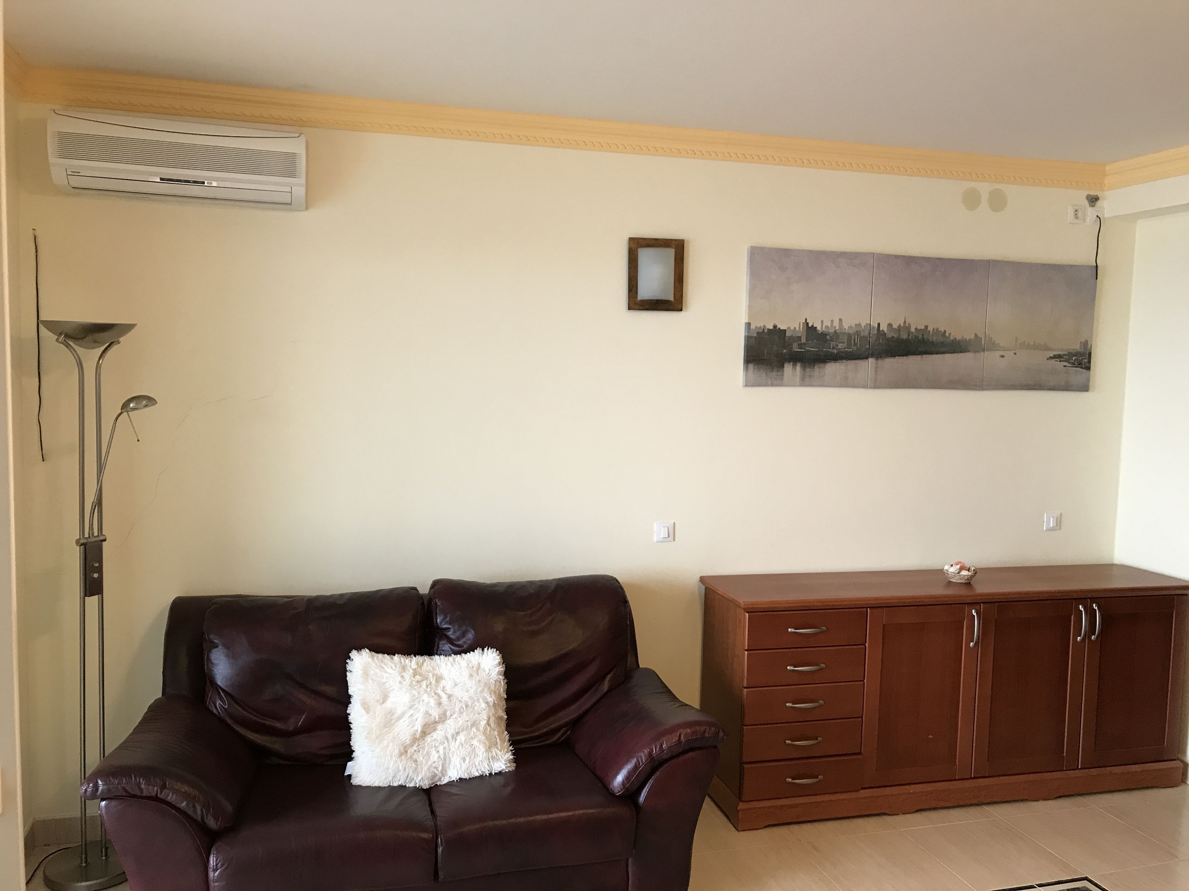 Una sala de estar con un sofá de cuero marrón, un cojín blanco y mullido, un mueble de madera y un cuadro con una vista urbana.