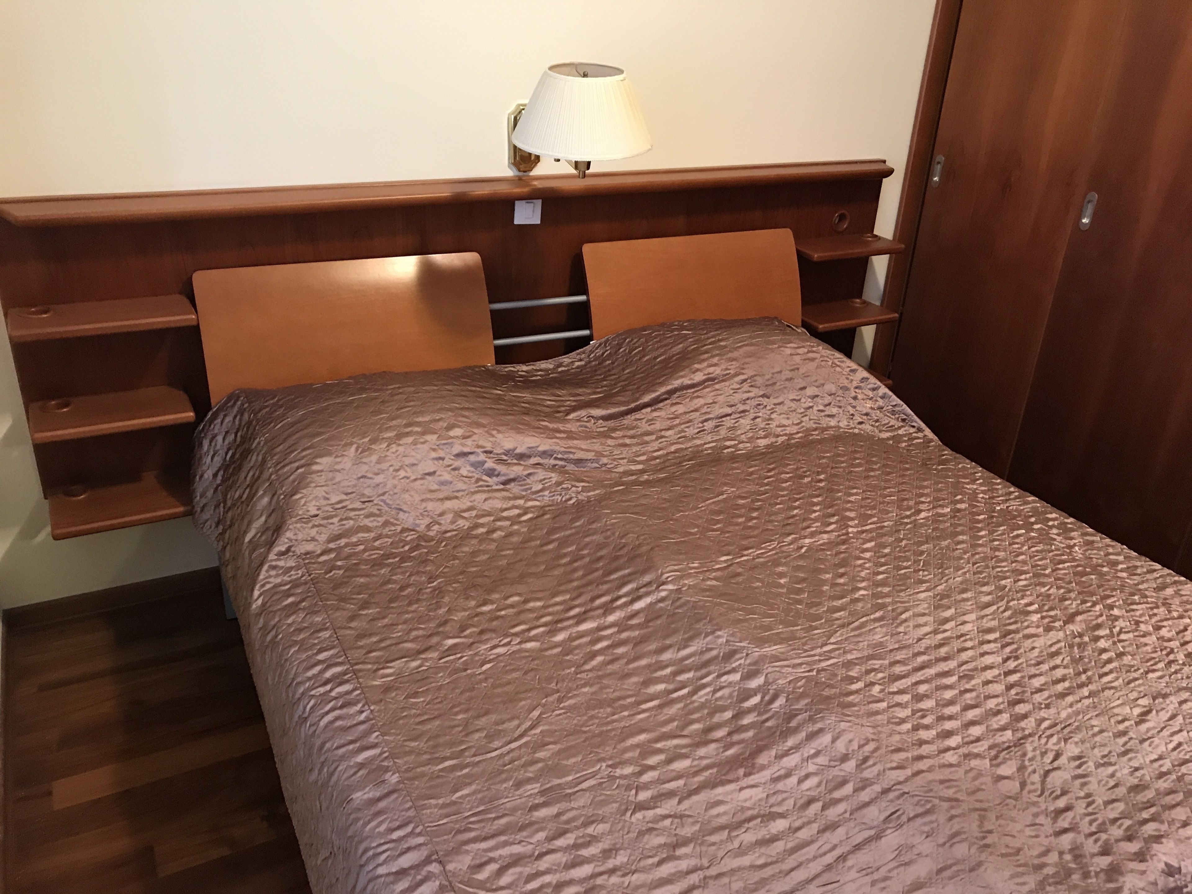 Un letto rifatto con cura, con copriletto marrone texturizzato, una testiera in legno con mensole integrate e una lampada fissata al muro.