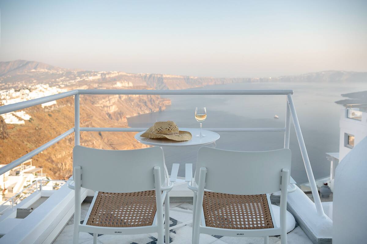 A balcony with two white chairs and a table, a straw hat, and a glass of wine.