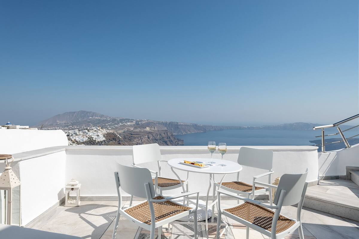 A terrace with white furniture, a table with a plate and glasses, and a scenic view of a coastline.