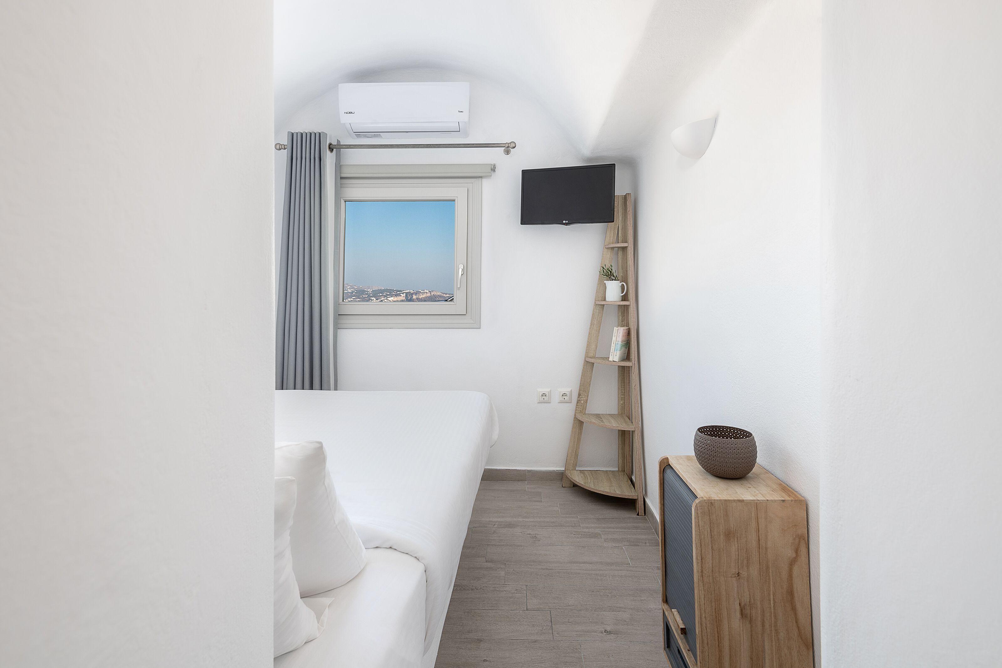 A hotel room with a bed, a TV mounted on the wall, a window with a view, and a wooden shelf with a bowl.