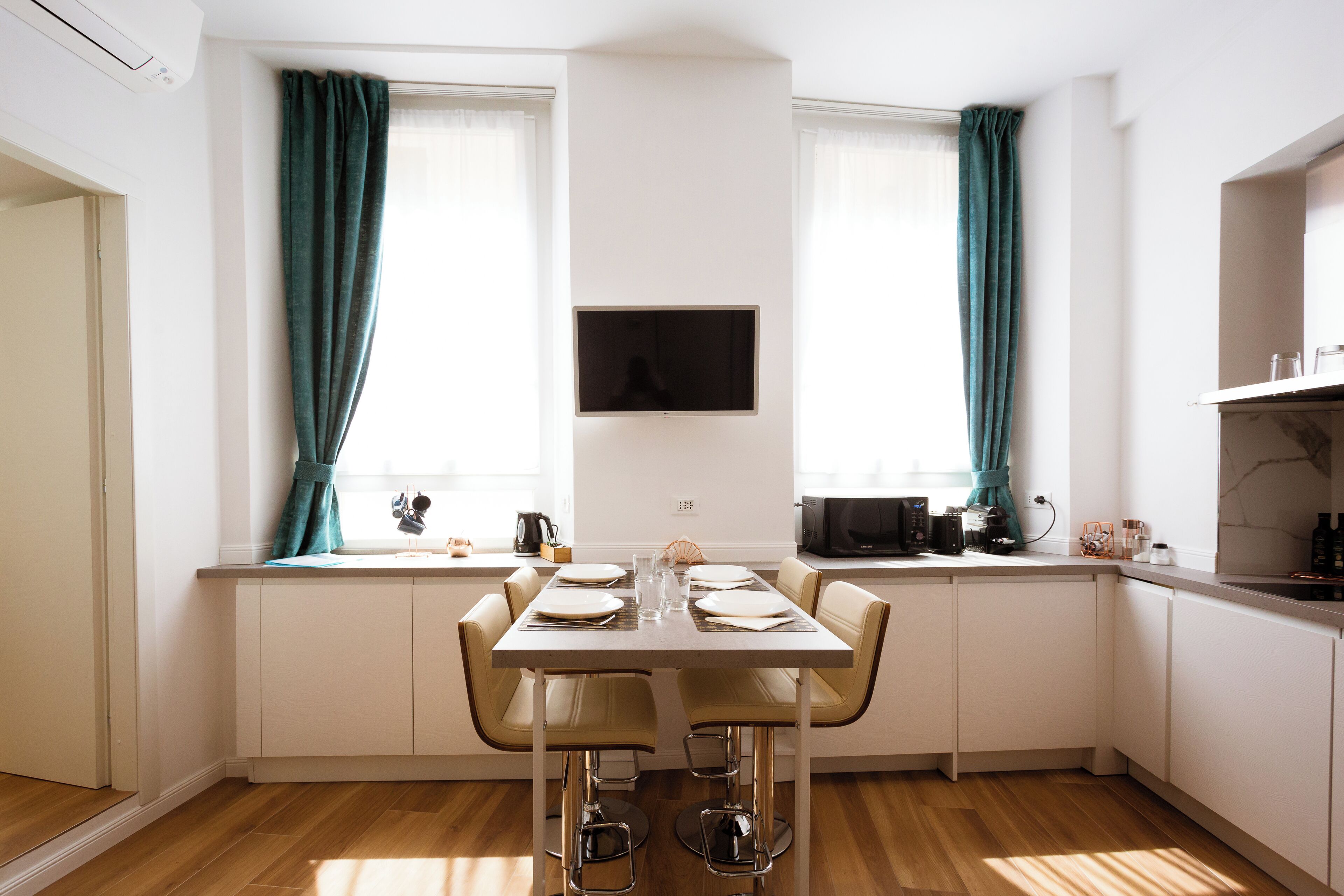 A modern kitchen with a dining table set for four, white cabinetry, teal curtains, and a flat-screen TV mounted above the counter.