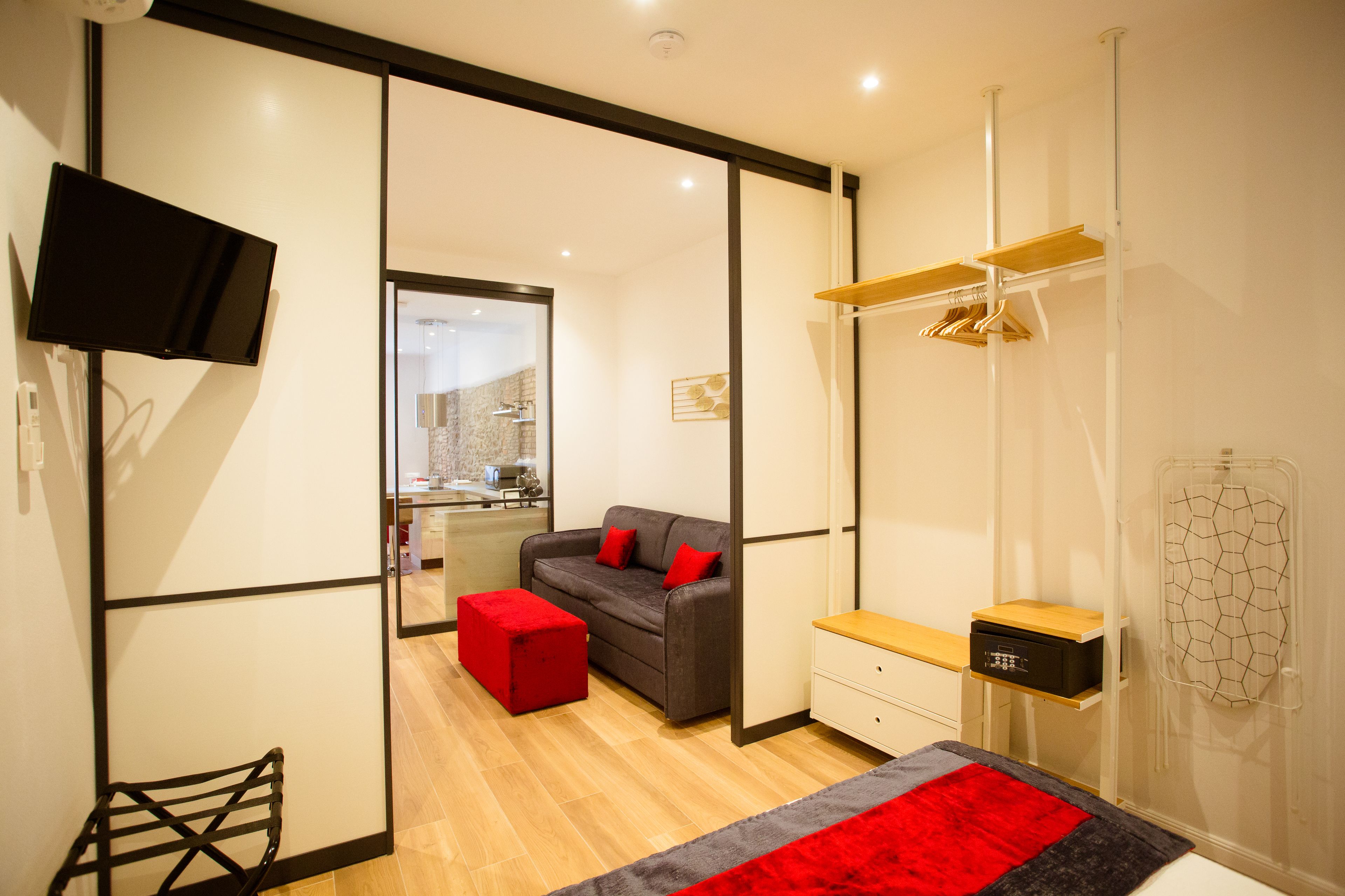 Een moderne slaapkamer met een grijze bank, een rode poef en een aan de muur bevestigde televisie.