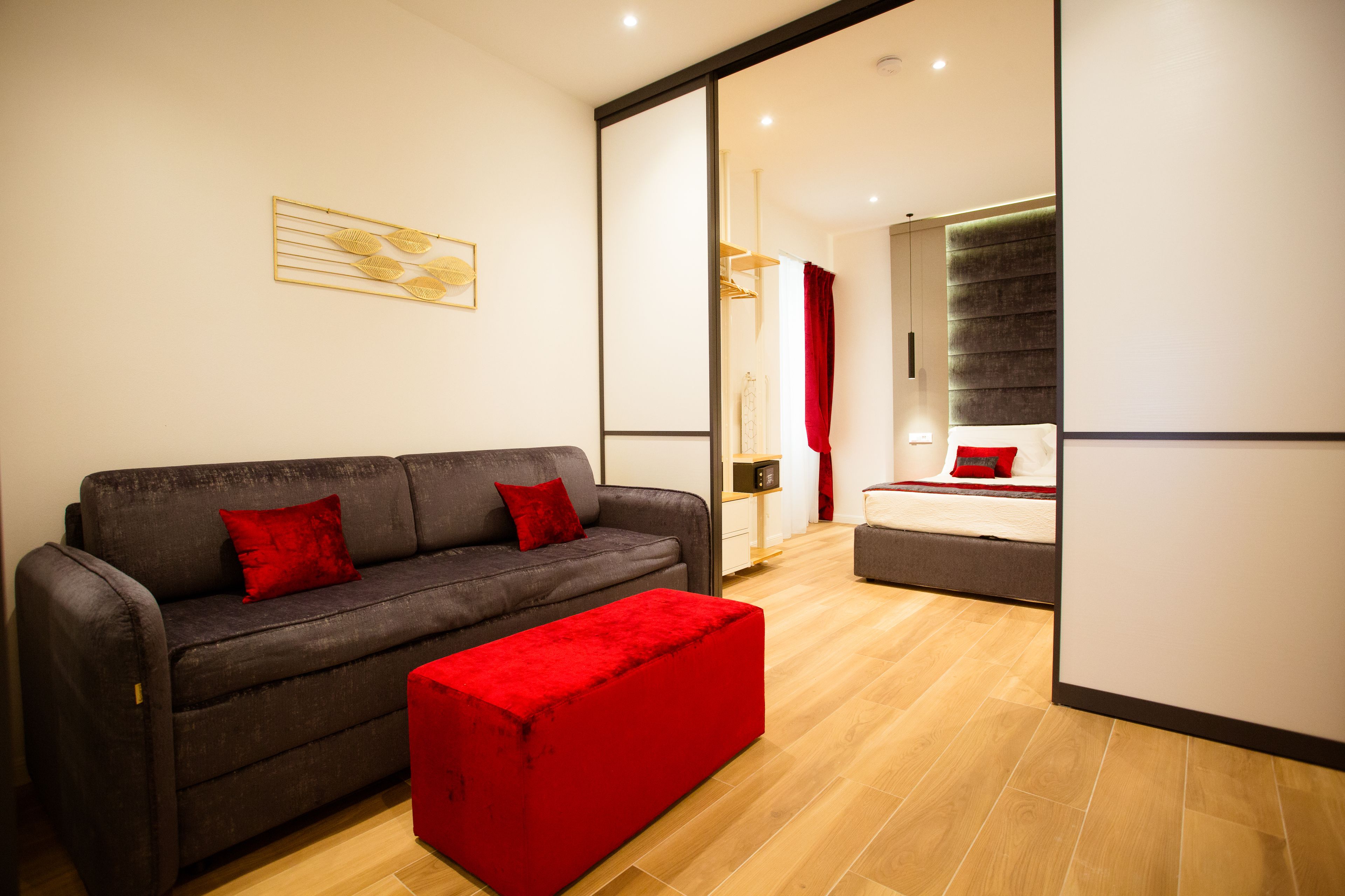 Un salon moderne avec un canapé gris, des coussins rouges et un pouf rouge. Un grand miroir reflète la pièce, et on peut voir un plancher en bois.