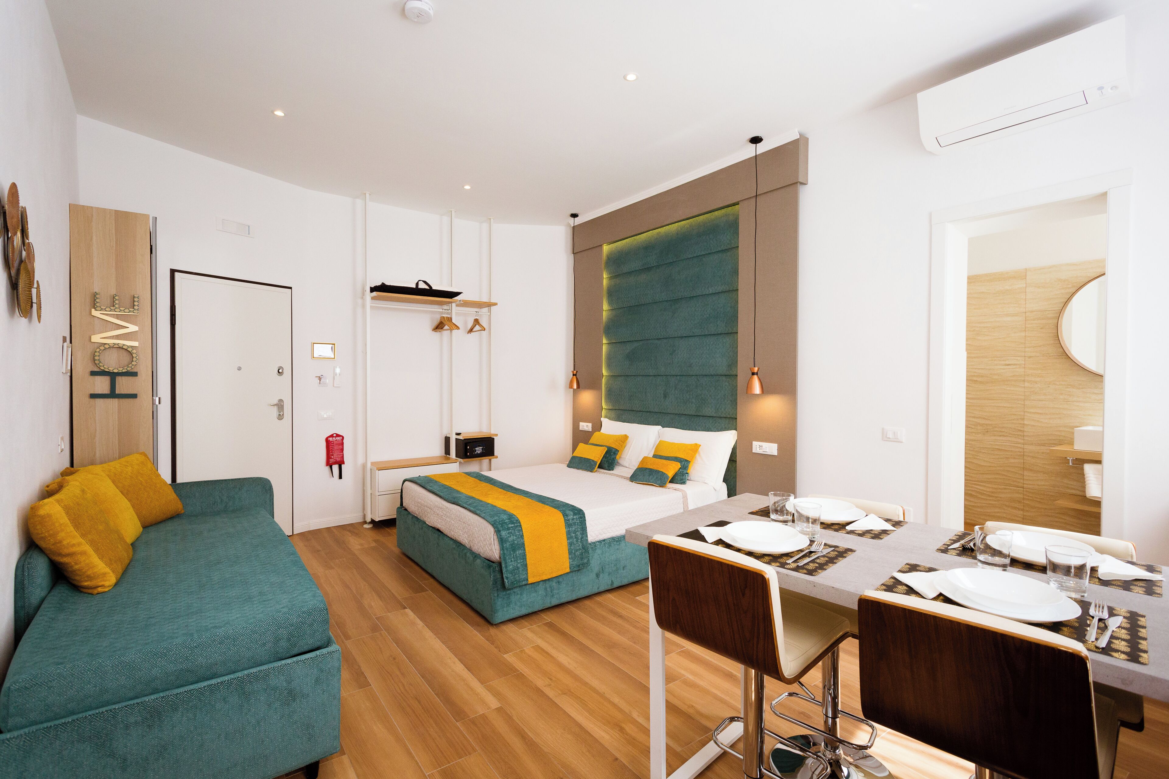 Une chambre d’hôtel moderne, équipée d’un lit, d’un canapé, d’une table à manger pour deux personnes et d’une salle de bain visible à travers une porte ouverte.