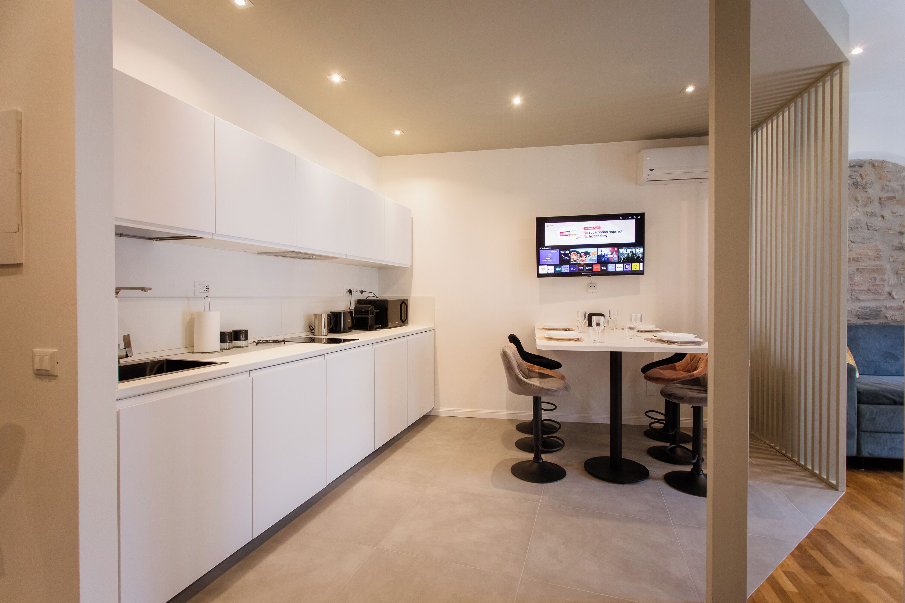 A modern kitchen with white cabinets, a dining area with a table and chairs, and a mounted TV displaying a news channel.