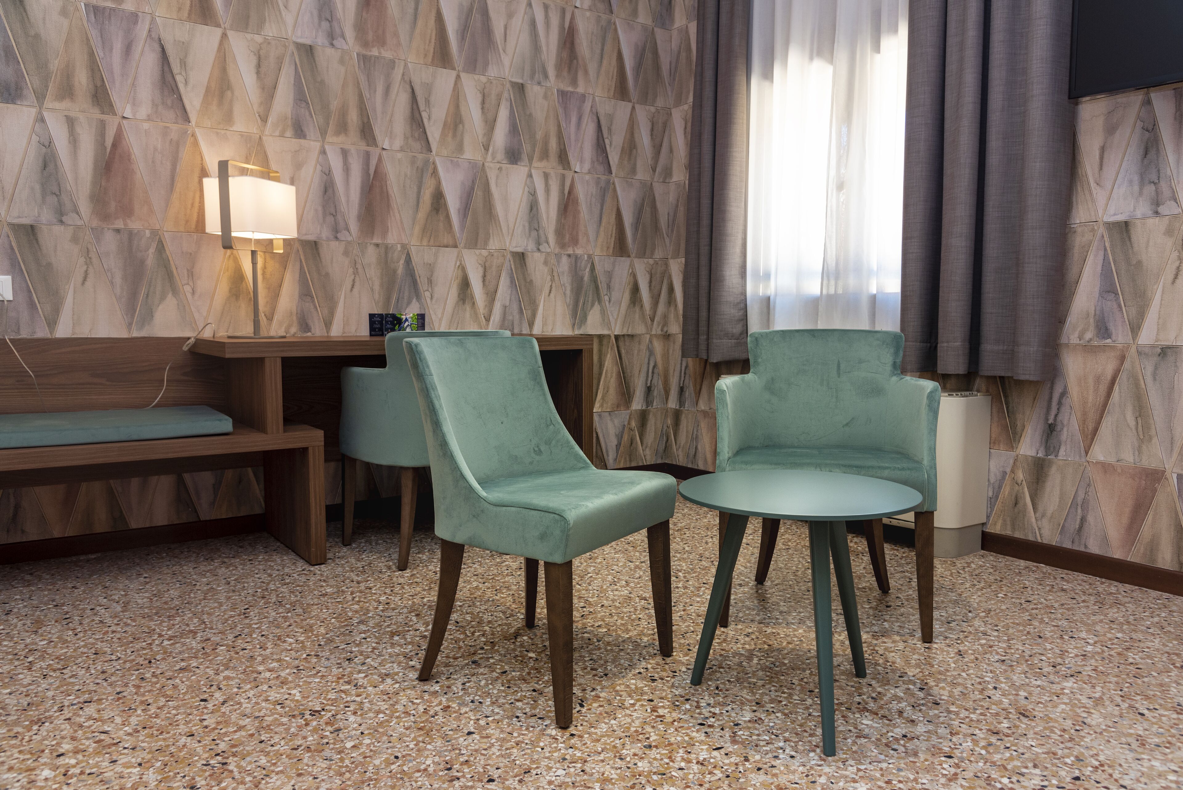A hotel room with a wooden bench, two green upholstered chairs, a small table, and a wall-mounted lamp.