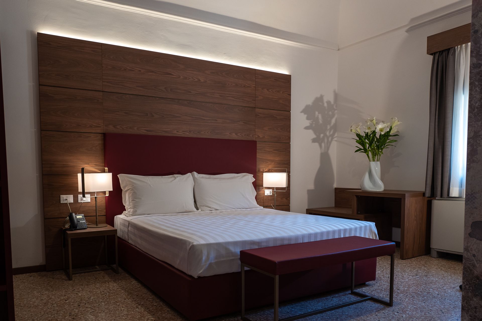 Ein Hotelzimmer mit einem großen Bett, Nachttischlampen, einem Telefon am Nachttisch, einem kleinen Tisch und einer Vase mit Blumen.