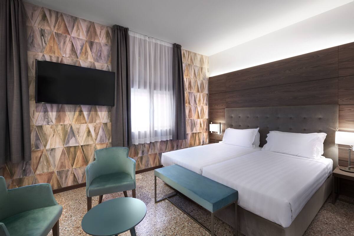 A hotel room with a large bed, two chairs, a bench, a TV mounted on a geometric-patterned wall, and a nightstand with a lamp.
