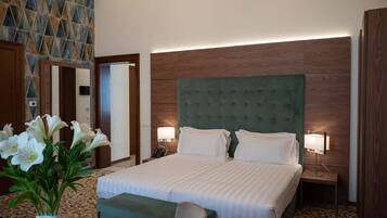 Une chambre d’hôtel avec un grand lit, deux tables de chevet avec des lampes, une tête de lit verte en tissu, un mur d’accent en bois et un vase rempli de fleurs blanches posé sur le sol.