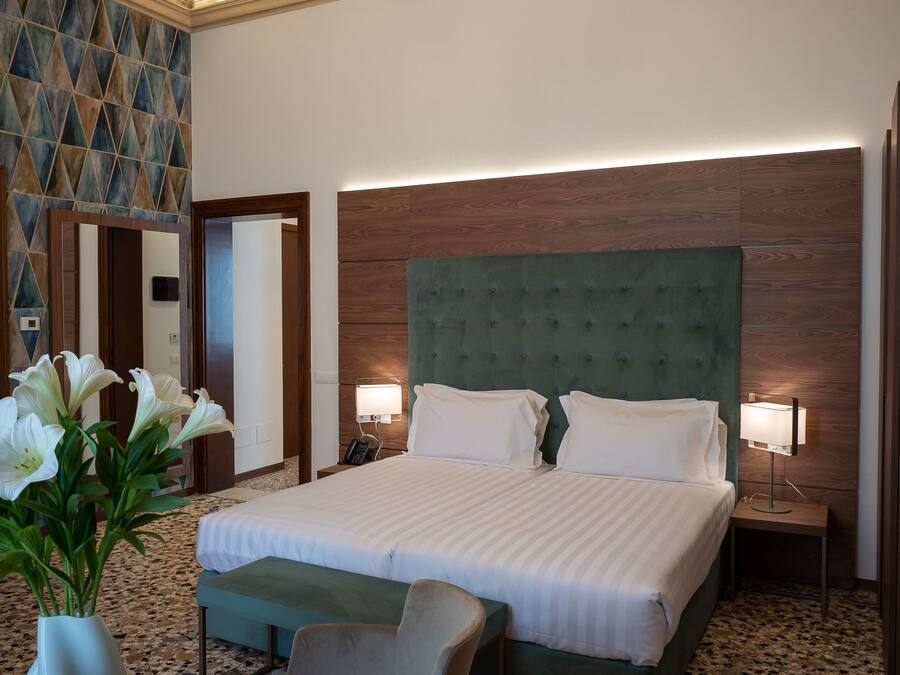 Une chambre d’hôtel avec un grand lit, deux tables de chevet avec des lampes, une tête de lit verte en tissu, un mur d’accent en bois et un vase rempli de fleurs blanches posé sur le sol.
