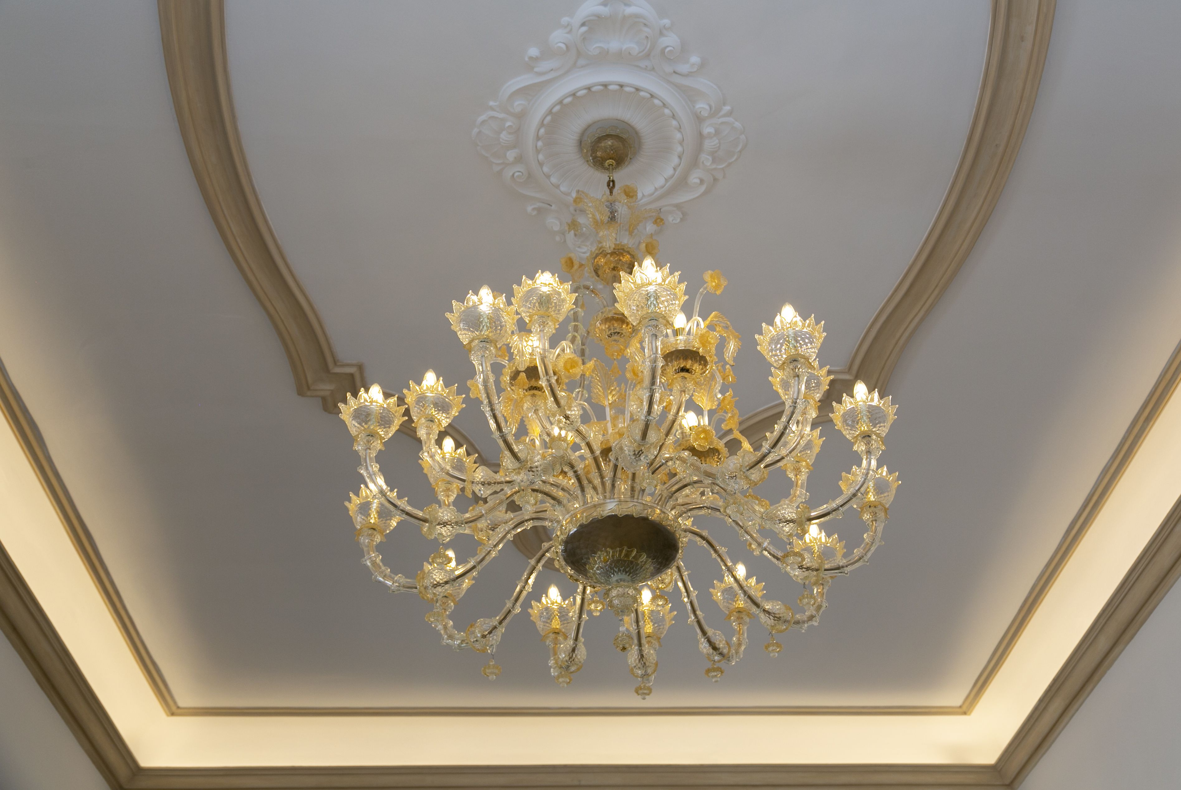 Un lustre à plusieurs bras, orné de lampes en forme de bougies, est installé dans une pièce dont le plafond est décoré de moulures travaillées.