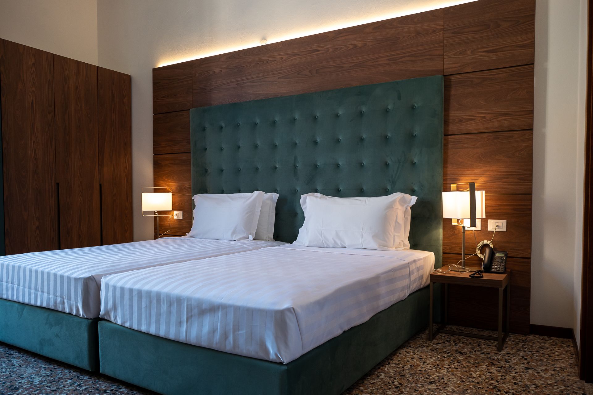 A double bed with a green upholstered headboard, two bedside lamps, and a small bedside table with a phone and charger.