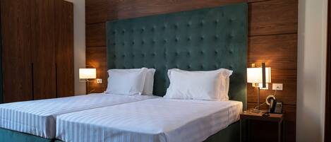 Una cama doble con cabecera tapizada de verde, dos lámparas de noche y una mesita de noche con un teléfono y cargador.