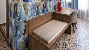 A hotel room with a geometric patterned wall, a flat-screen TV, a red vase, a wooden desk, a cushioned bench, and a terrazzo floor. - Palazzo Scamozzi (Vicenza)