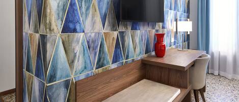 A hotel room with a geometric patterned wall, a flat-screen TV, a red vase, a wooden desk, a cushioned bench, and a terrazzo floor.