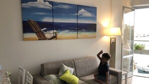 Een woonkamer met een beige bank, een bijzettafeltje en een groot schilderij met een strandthema aan de muur.