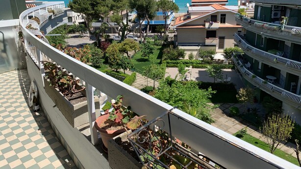 Een balkon met een geruite vloer, potplanten en uitzicht op een kustgebied met gebouwen en bomen.