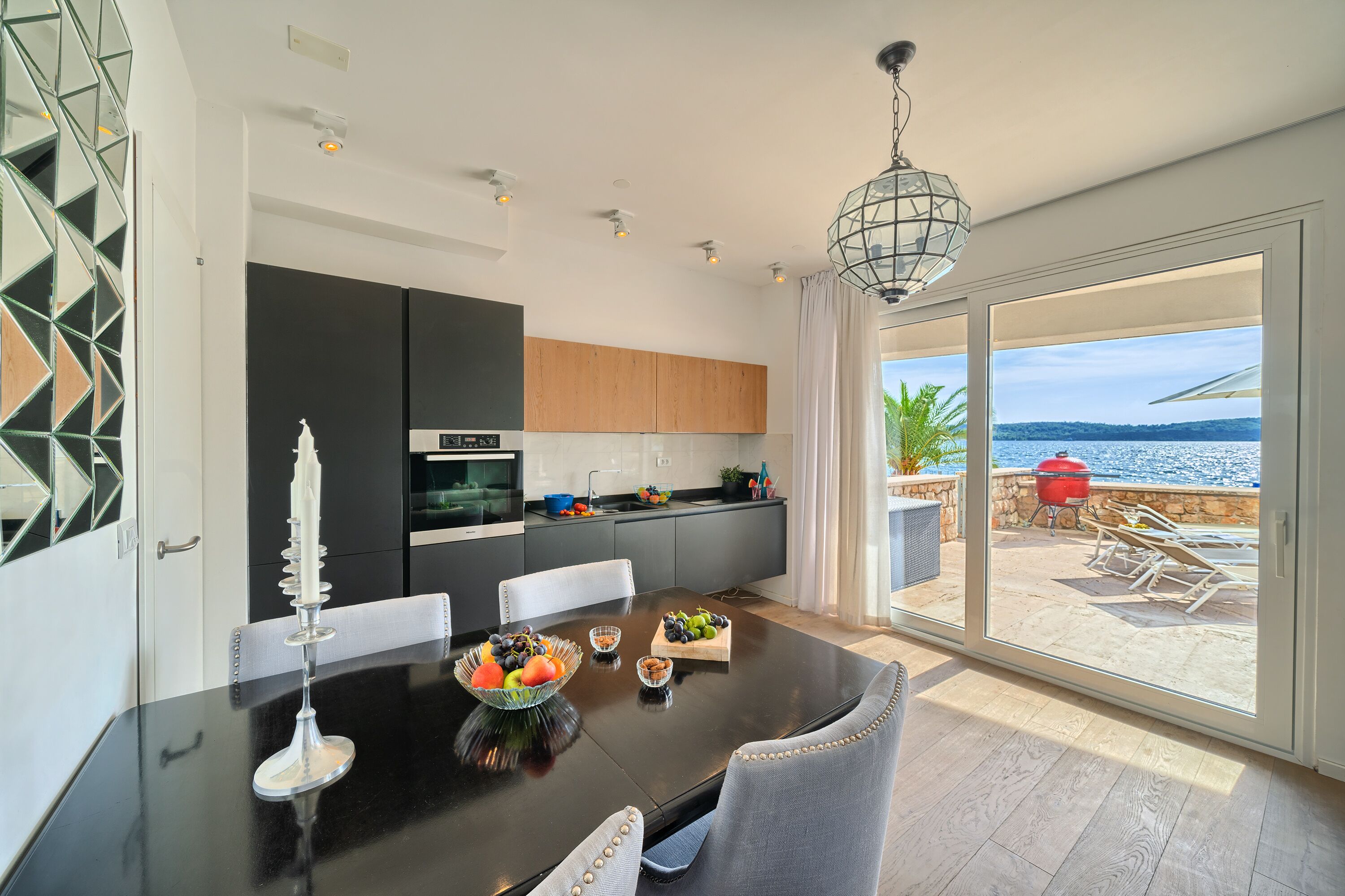 A modern kitchen with dark cabinets, a dining table with fruit, and a view of the sea through large sliding doors.