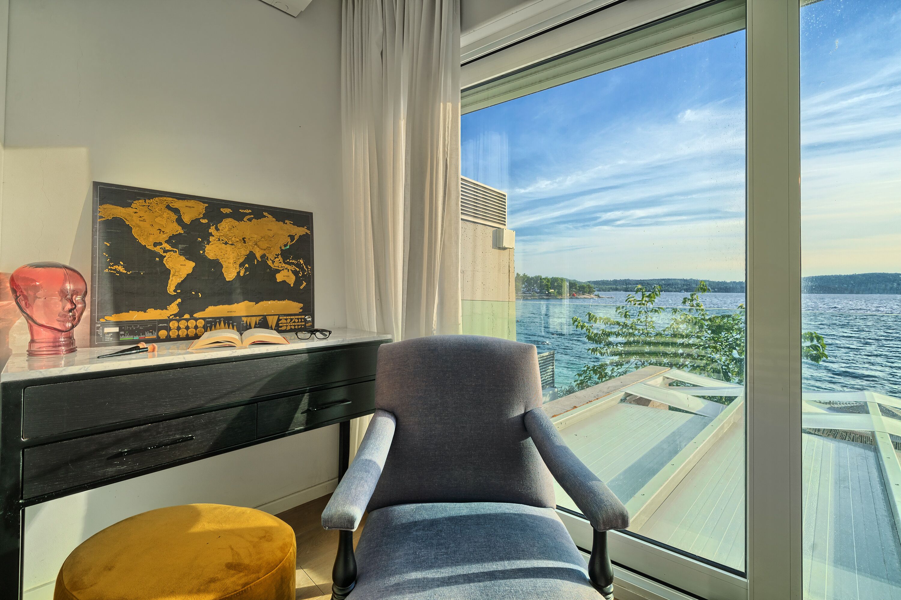 Una habitación con escritorio, silla, un cuadro de un mapa mundial y vistas a un cuerpo de agua a través de un ventanal grande.