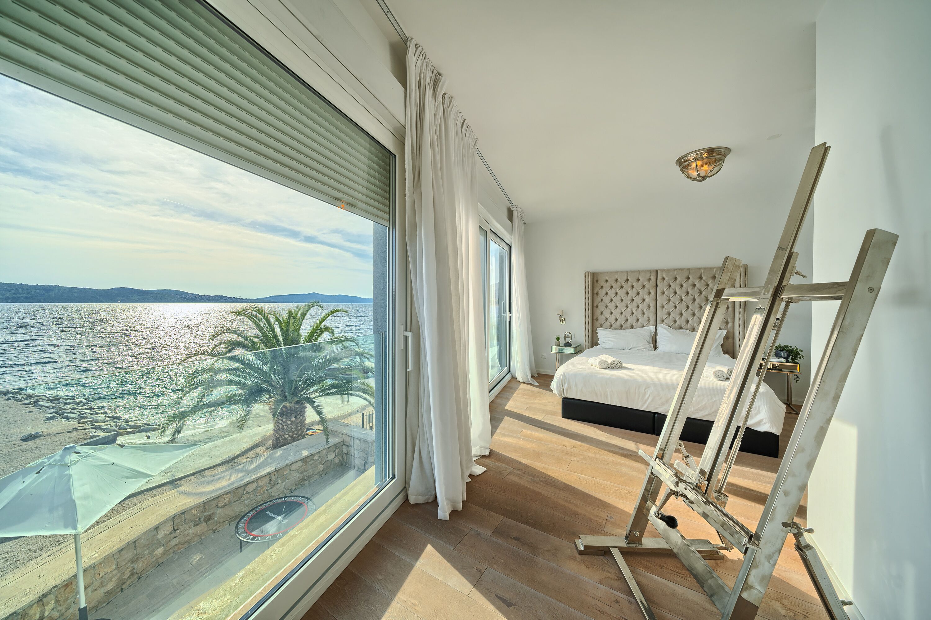 A bedroom with a large window overlooking a beach, a bed, and an easel.