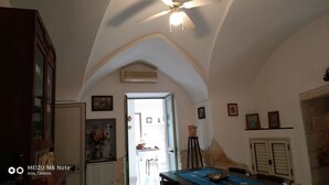 Une pièce avec un plafond haut, une porte en arc de cercle, un ventilateur de plafond, une table à manger recouverte d’une nappe bleue, et une cuisine visible à travers l’ouverture.