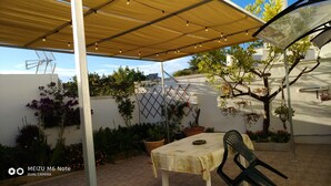 Une terrasse extérieure avec une table et des chaises, une pergola illuminée de guirlandes lumineuses, des plantes en pot et une plante grimpante.