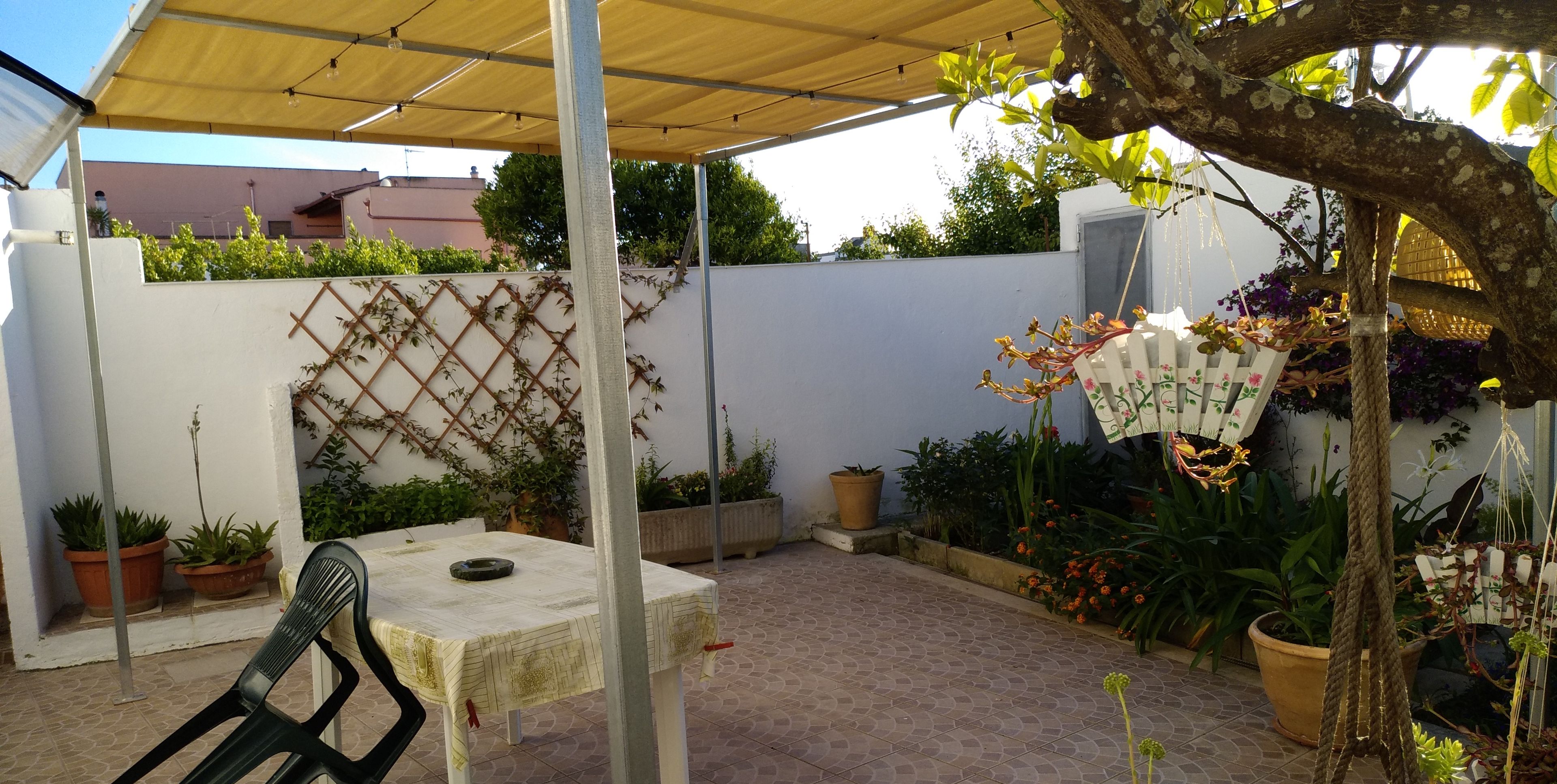 Een overdekt terras met een tafel en stoelen, omringd door potplanten en een decoratieve ventilator.
